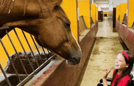 Carolina, en fauteuil roulant, salue un cheval qui passe la tête hors de son box.