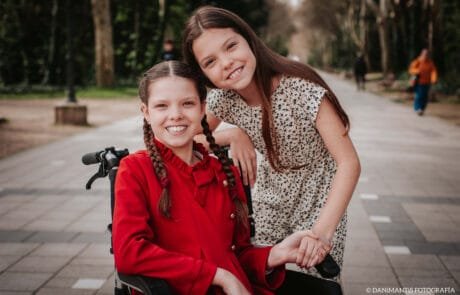 Deux sœurs sourient dans un parc ; l’aînée, vêtue d’un manteau rouge, est en fauteuil roulant.