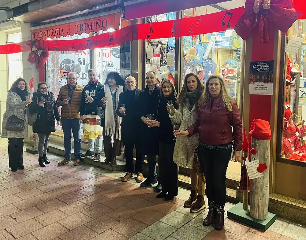 Un groupe trinque devant un commerce décoré de rubans rouges et de notes de musique, près du panneau du concours de Noël.