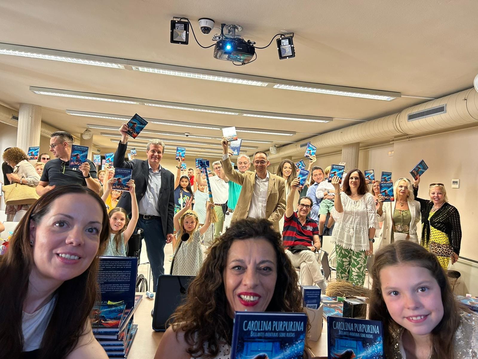 Selfie lors de la présentation : le public brandit des exemplaires de « Carolina Purpurina » au-dessus d’une table de livres.