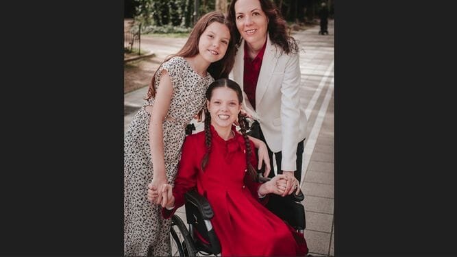Portrait en extérieur : Carolina, en rouge dans son fauteuil roulant, tient la main de sa sœur et de leur mère.