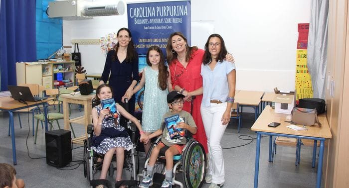 Photo de groupe en classe devant le kakémono : trois femmes, une fille et deux enfants en fauteuil roulant avec le livre.