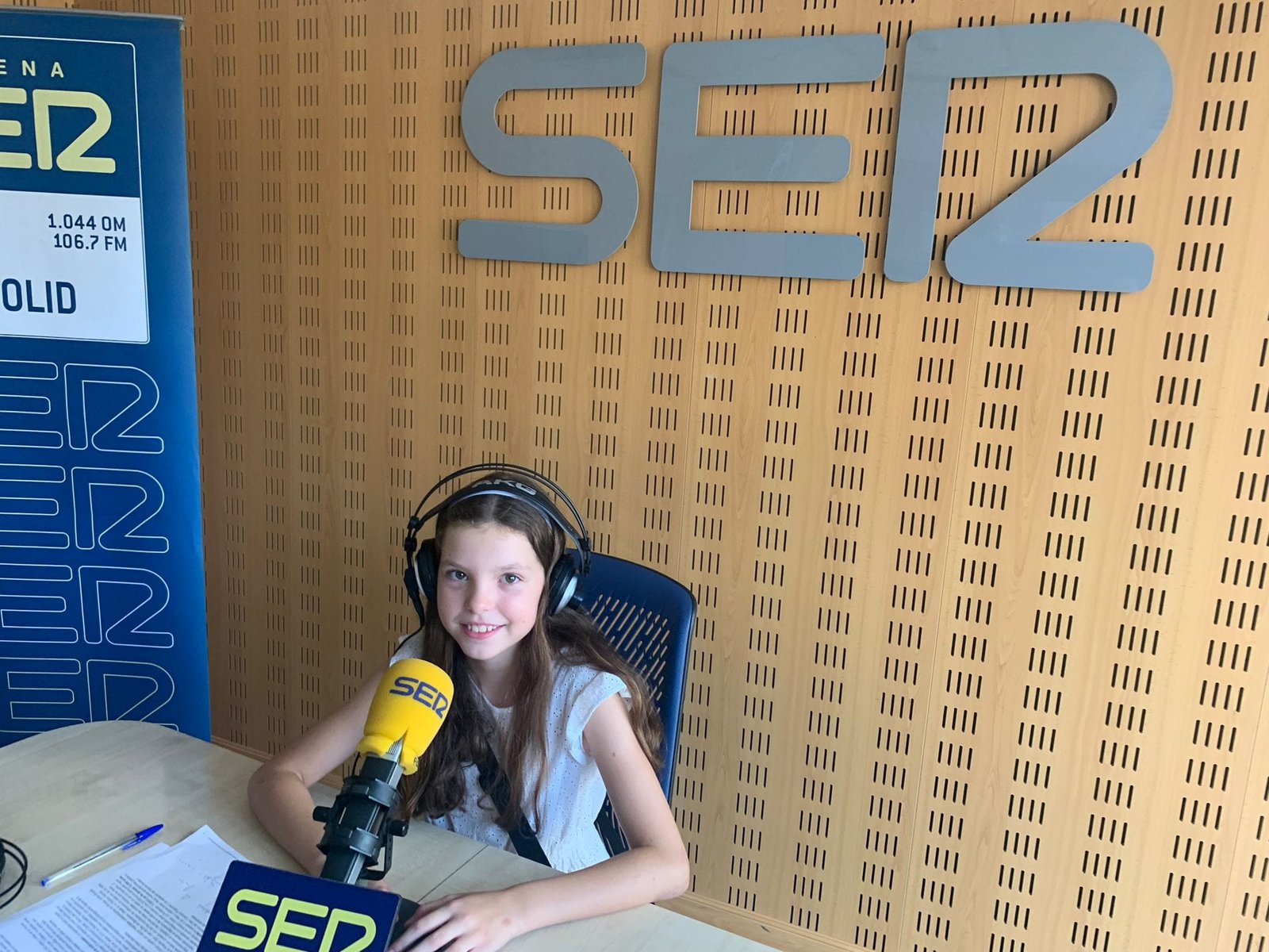 Une fille au casque sourit devant le micro jaune de la Cadena SER, sous le logo de la station dans le studio.