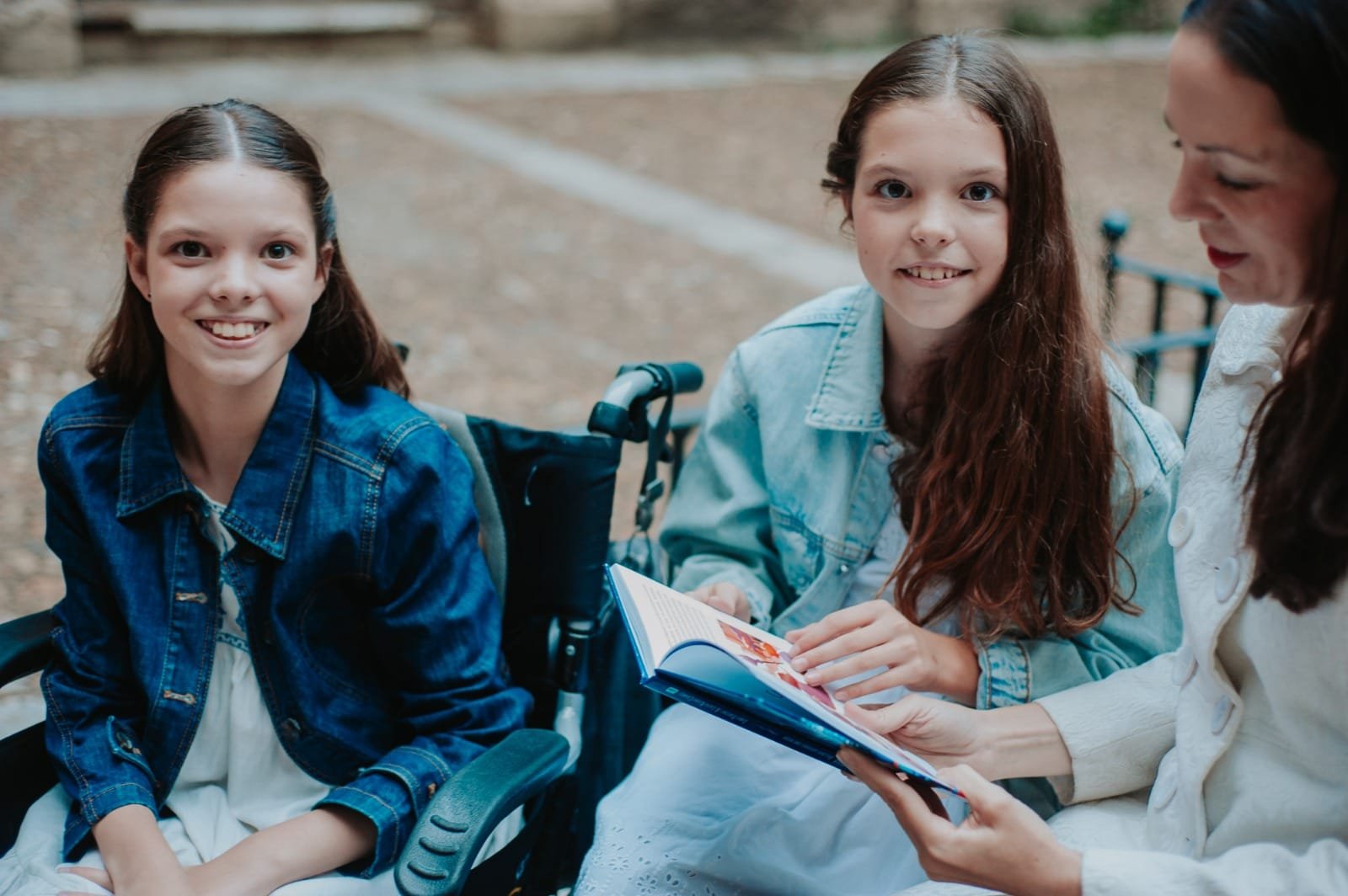 En extérieur, une fille en fauteuil roulant à côté d’une autre fille et d’une femme qui feuillettent le livre ouvert.