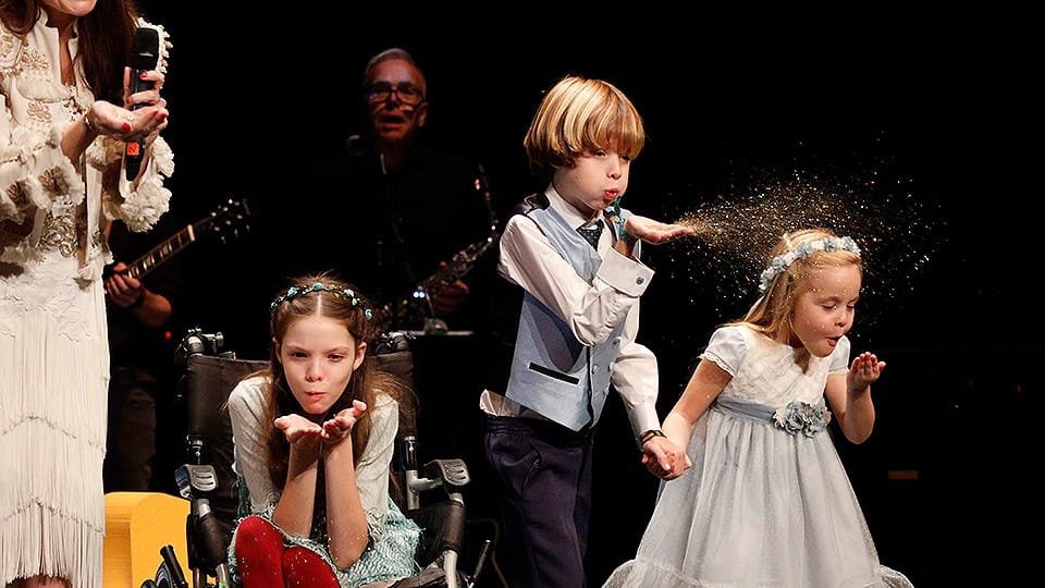 Sur scène, trois enfants soufflent des paillettes ; l’une d’elles, en fauteuil roulant, porte une couronne de fleurs.