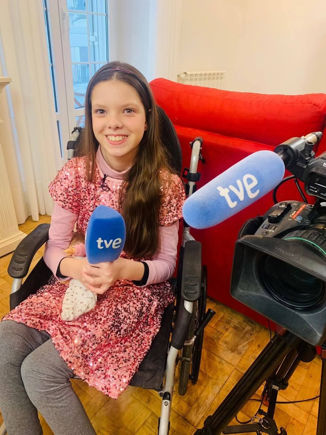 Carolina, dans son fauteuil roulant et en robe rose à paillettes, tient un micro de TVE devant la caméra.