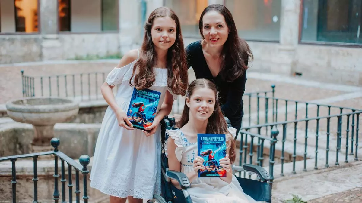 Dans un patio, une fille en fauteuil roulant et une autre debout montrent la couverture de « Carolina Purpurina », une femme à leurs côtés.