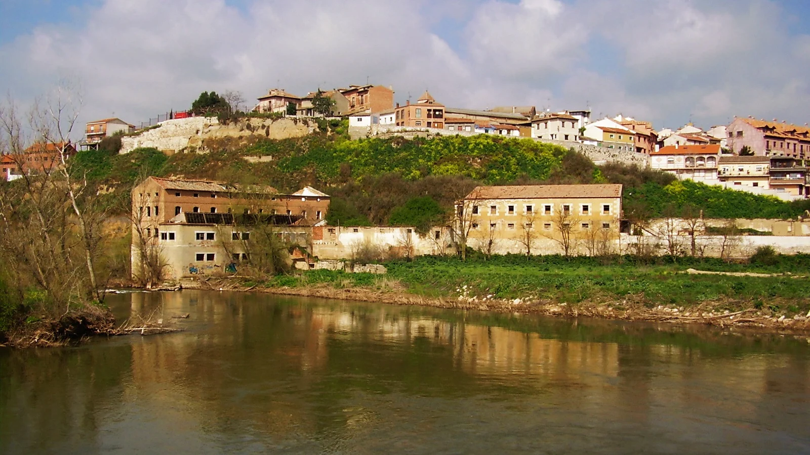Vue d’un village perché sur une colline au bord d’une rivière, avec de vieux bâtiments sur la rive.