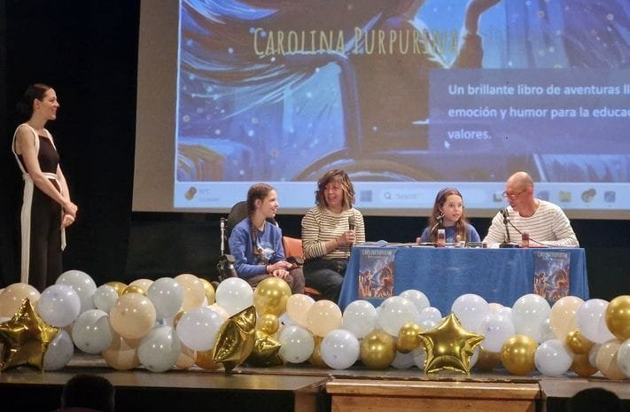 Rencontre sur la scène d’une école : quatre personnes à la table, l’une au micro, au milieu de ballons dorés et blancs.