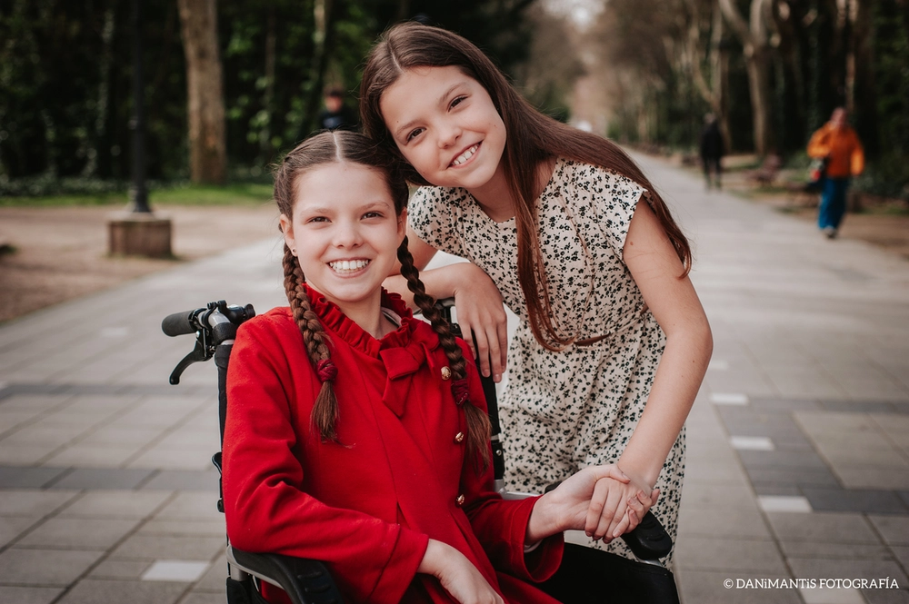 Carolina, en fauteuil roulant et manteau rouge, sourit aux côtés d’une autre fillette qui l’enlace dans une allée d’arbres.