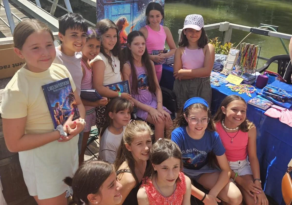 Un groupe de fillettes pose près du stand de vente au bord de la rivière ; plusieurs tiennent le livre « Carolina Purpurina ».