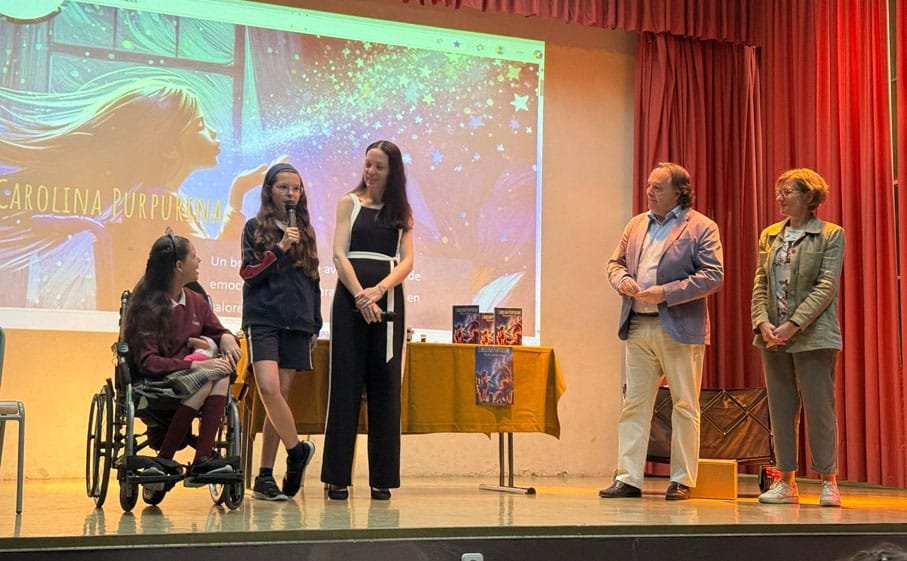 Sur scène, une adolescente parle au micro auprès de Carolina en fauteuil roulant, la couverture du livre projetée derrière elles.