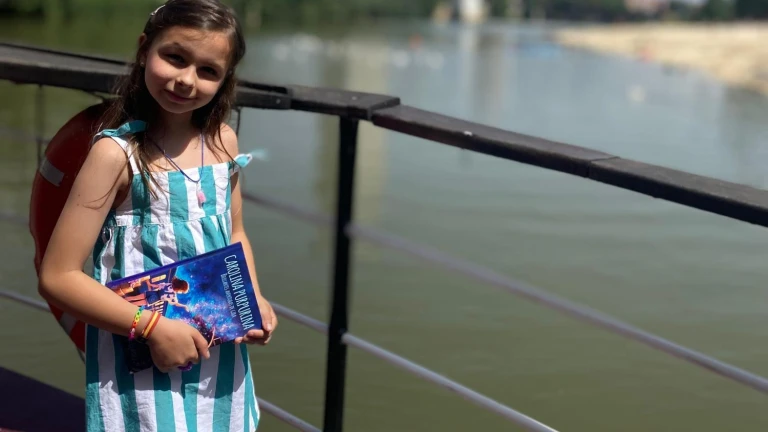 Une fillette en robe rayée, sac au dos, tient un exemplaire de « Carolina Purpurina » près du garde-corps au bord d’une rivière.