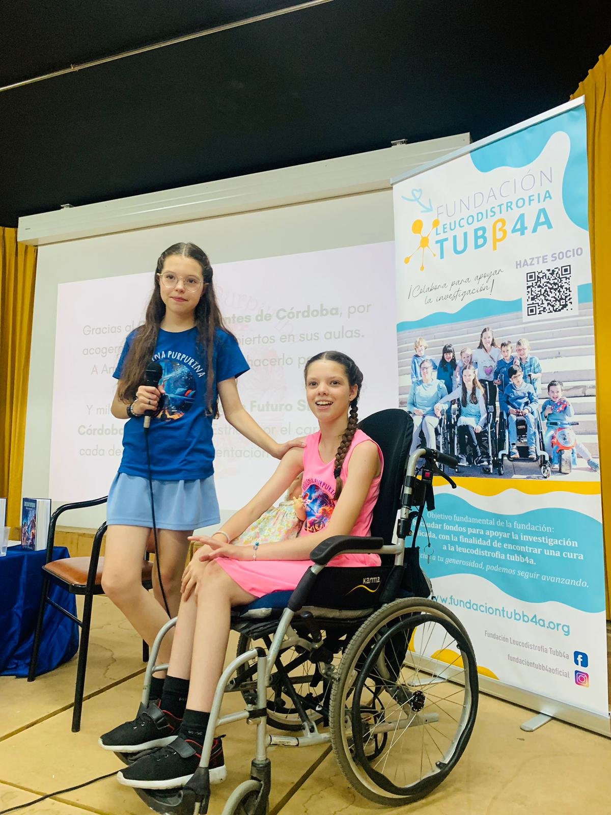 Isabel, micro à la main, aux côtés de Carolina en fauteuil roulant, devant le kakémono de la Fundación Leucodistrofia TUBB4A.