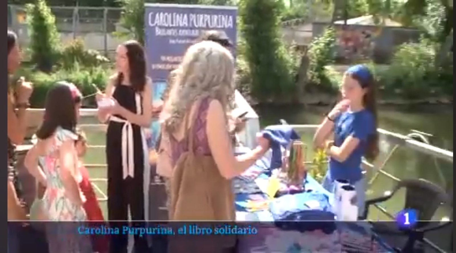 Image du reportage de TVE titrée « Carolina Purpurina, el libro solidario » : des visiteurs devant le stand du livre au bord de l’eau.