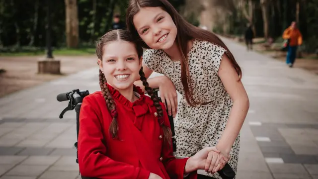 Carolina, dans son fauteuil roulant, sourit tandis qu’une autre fille s’appuie sur son épaule dans une allée arborée.