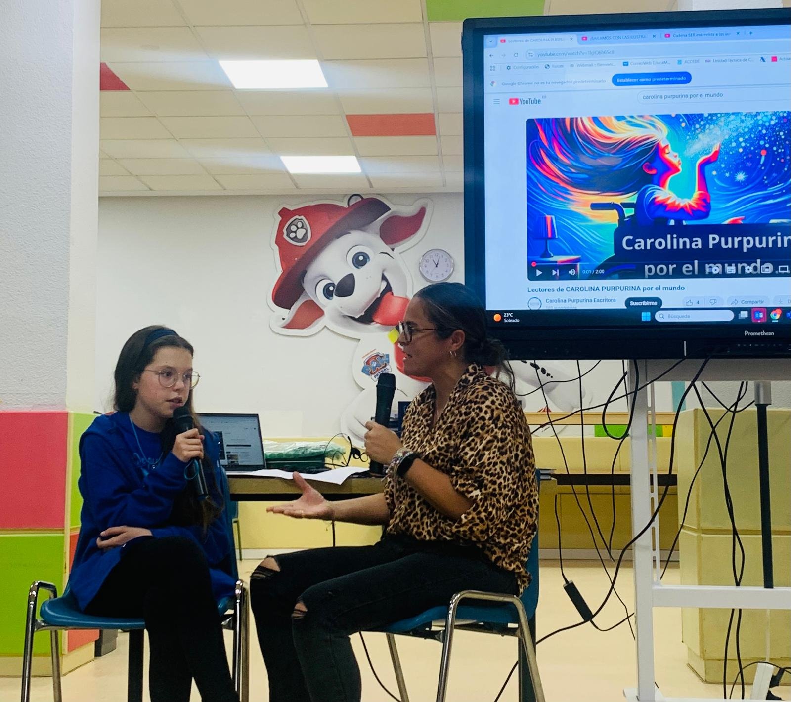 Isabel, micro en main, répond à une intervieweuse devant un écran qui diffuse la vidéo « Carolina Purpurina por el mundo ».