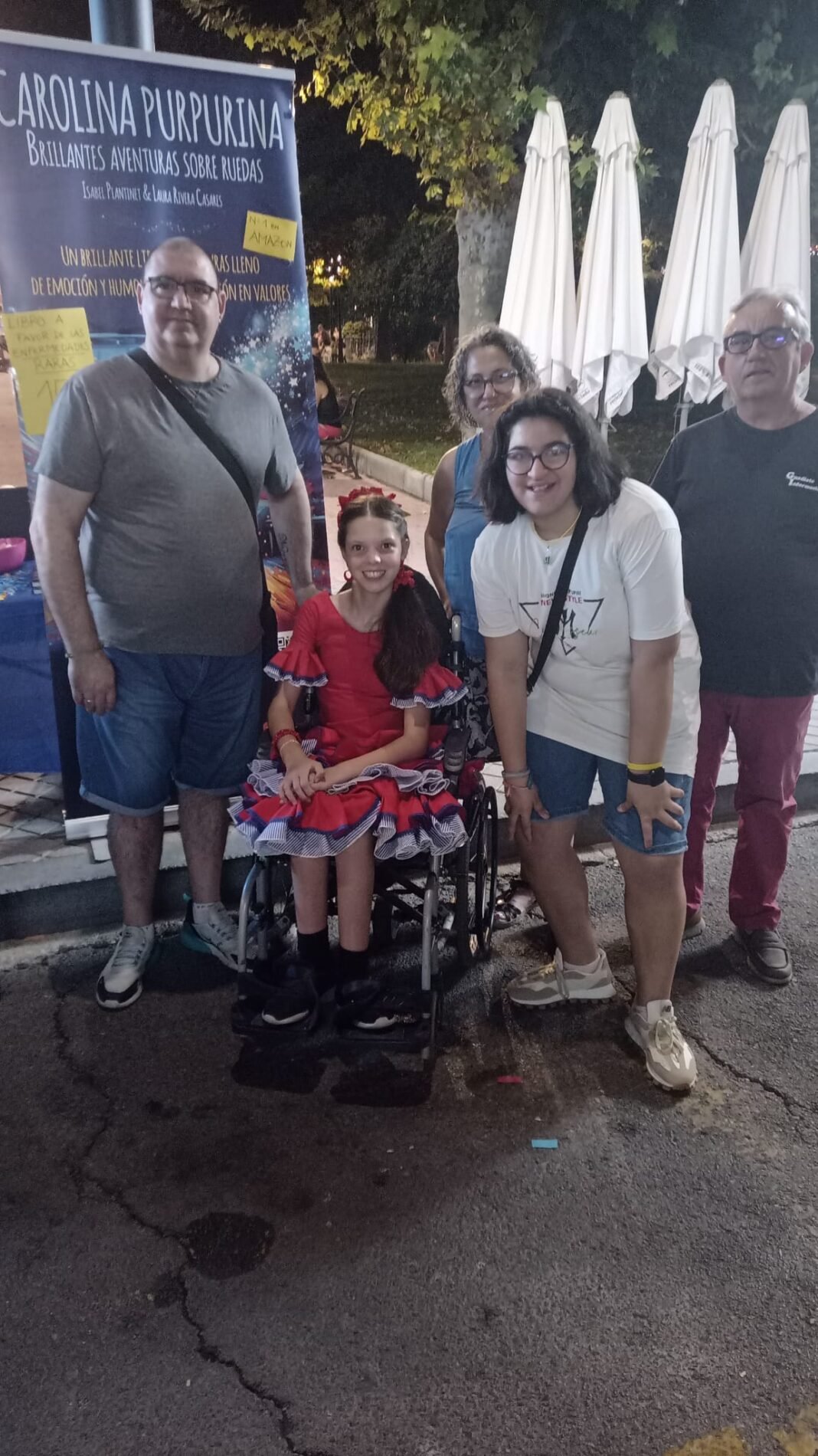 Le soir à la feria, une fille en robe de flamenca dans son fauteuil roulant pose avec plusieurs personnes devant le kakémono du livre.
