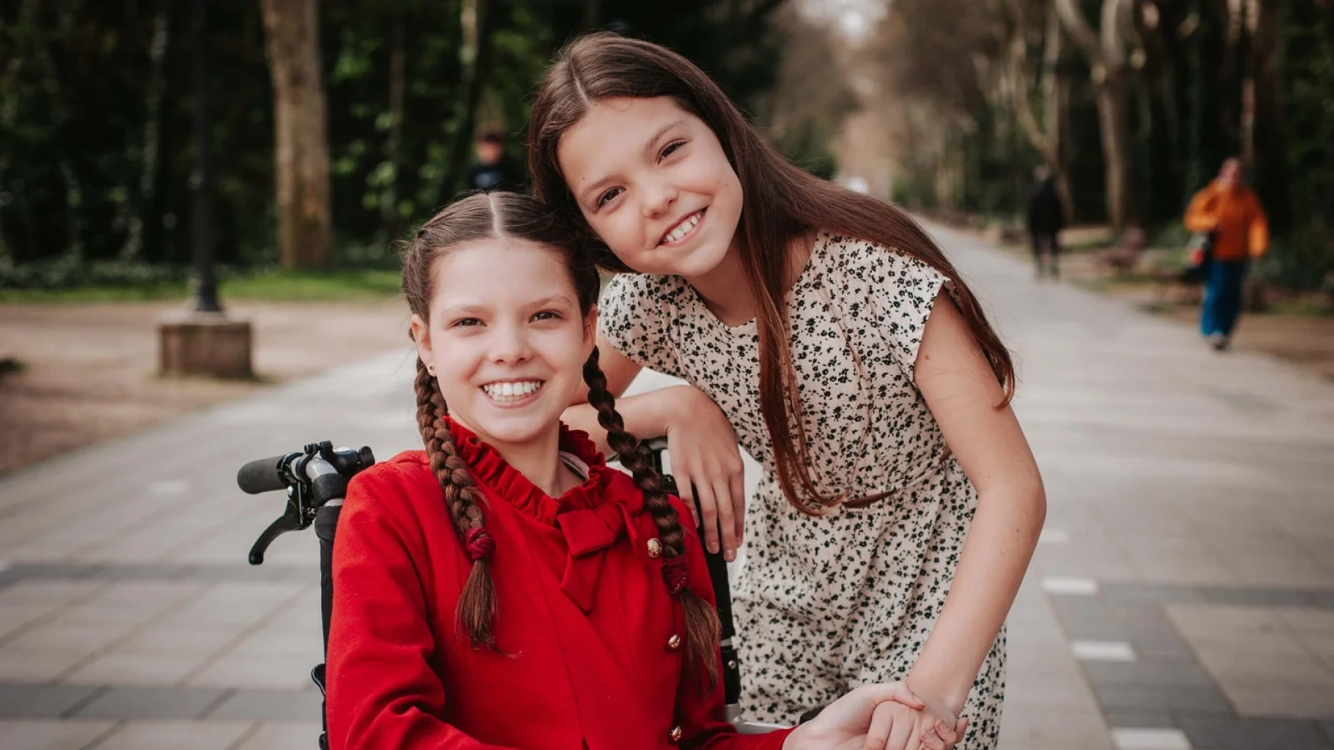 Carolina, en manteau rouge et nattes, sourit dans son fauteuil roulant près d’une autre fillette qui lui tient la main, dans une allée arborée.