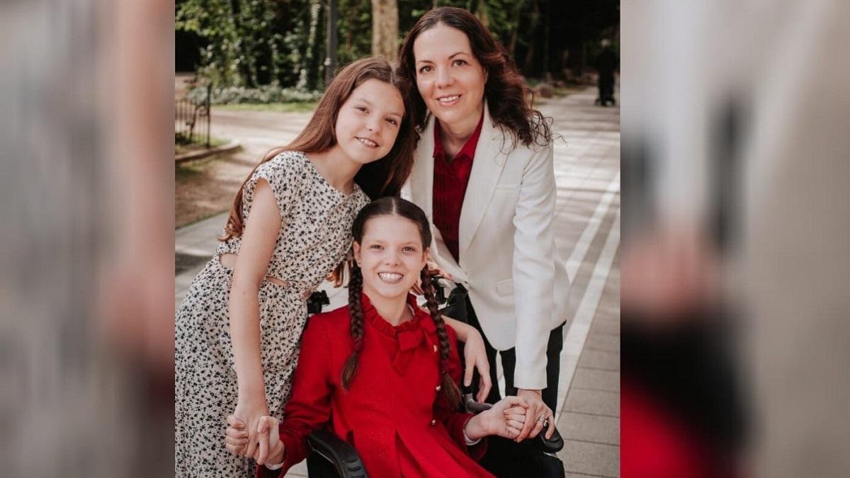 Carolina, en robe rouge et tresses, sourit dans son fauteuil roulant, entre une fillette en robe imprimée et une femme en veste blanche.