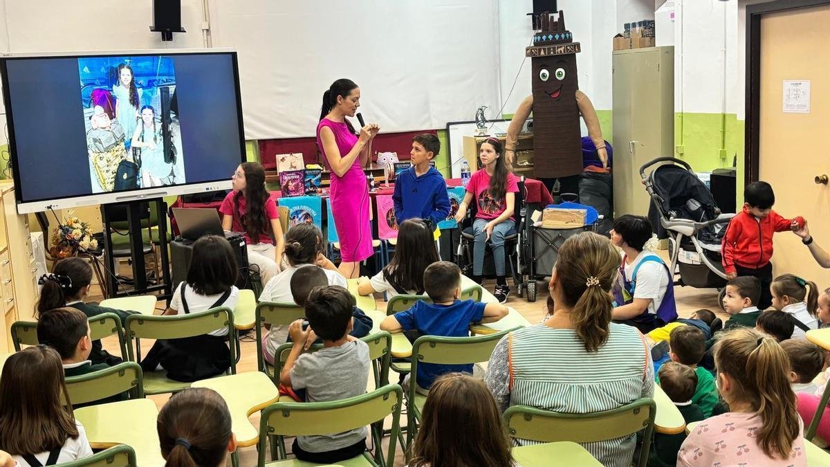Une femme parle au micro devant les élèves assis ; à ses côtés, des enfants et Carolina, dans son fauteuil roulant, près d’une table de livres.