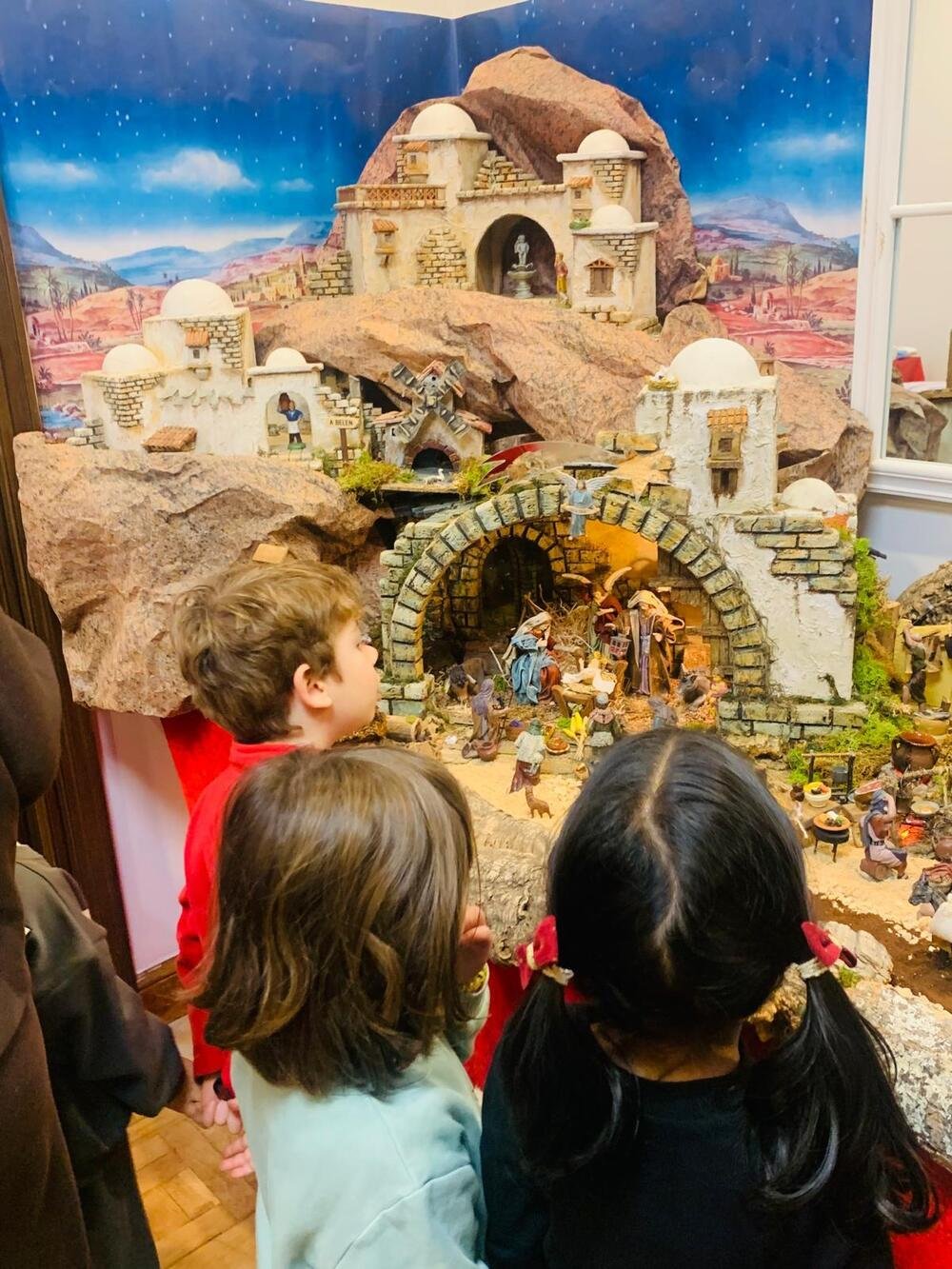 Trois enfants, de dos, contemplent la crèche : maisons blanches, une étable en pierre abritant la Nativité et un fond de ciel étoilé.