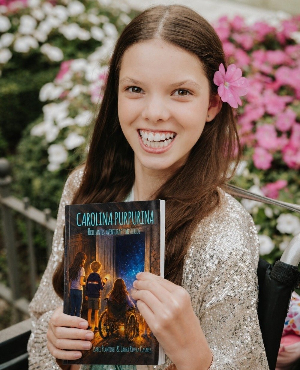 Une fillette souriante, une fleur rose dans les cheveux, tient le livre « Carolina Purpurina. Brillantes aventuras y misterios ».