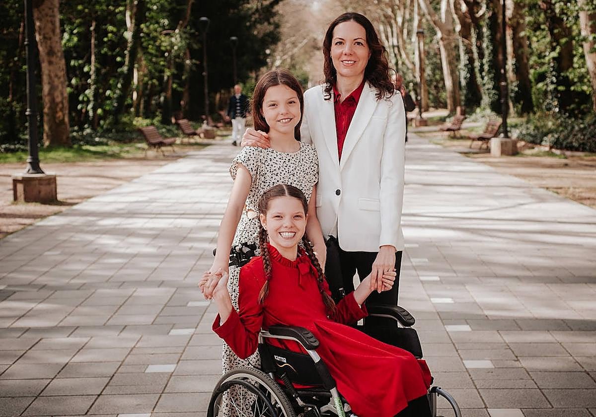 Carolina, dans son fauteuil roulant et en robe rouge, pose avec sa famille dans une allée arborée d’un parc.
