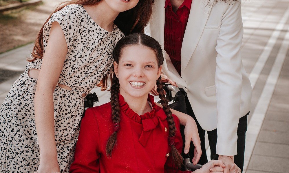 Carolina, en robe rouge et tresses, sourit dans son fauteuil roulant, entourée de sa famille, dont les visages sortent du cadre.