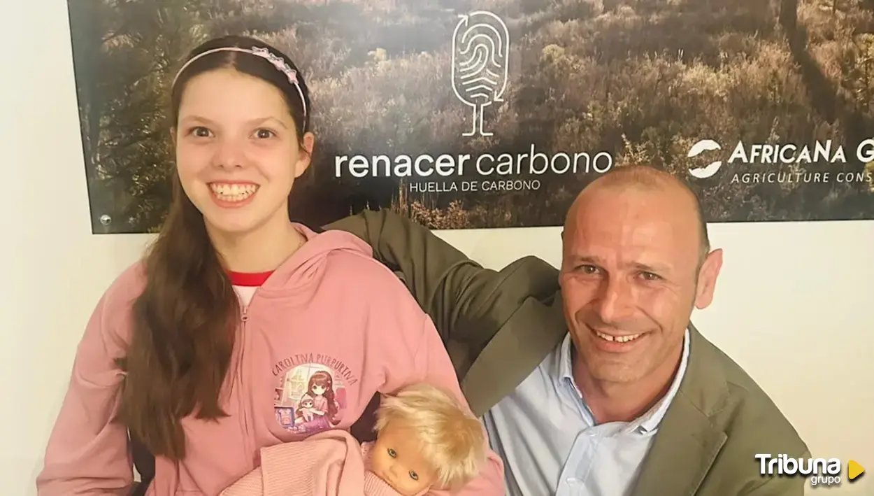 Une fillette en sweat rose Carolina Purpurina, une poupée dans les bras, pose avec un homme devant l’affiche de Renacer Carbono.